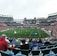 A large sports stadium filled with spectators. The field is green with white markings, and players in red and white uniforms are actively engaged in a game. The stands are packed with people wearing various colored clothing, and some sections of the crowd are more densely populated. Digital screens and advertisements are visible around the stadium, adding to the vibrant atmosphere.