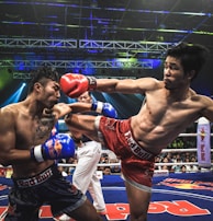 Two athletes are engaged in a dynamic kickboxing match inside a ring, surrounded by a cheering audience. One fighter, wearing red gloves and shorts, executes a high kick towards his opponent, who attempts to block the attack with his arm. The background features colorful lighting and promotional banners.