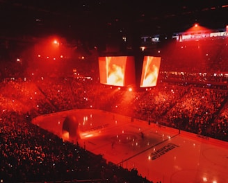 An indoor ice hockey arena filled with a large crowd, bathed in red lighting. A giant central scoreboard projects fiery visuals, illuminating the ice and audience. The atmosphere is charged, likely during a live sporting event.