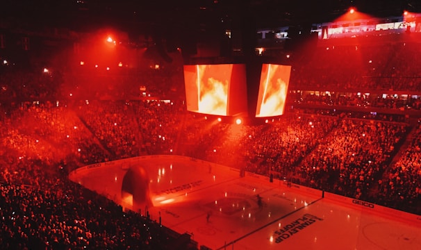 An indoor ice hockey arena filled with a large crowd, bathed in red lighting. A giant central scoreboard projects fiery visuals, illuminating the ice and audience. The atmosphere is charged, likely during a live sporting event.
