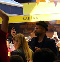 A lively crowd gathers in an outdoor setting with many people capturing the moment on their phones. The atmosphere is cheerful and energetic, with some individuals wearing sports jerseys, likely celebrating or supporting an event.