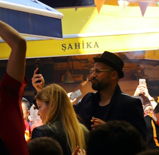 A lively crowd gathers in an outdoor setting with many people capturing the moment on their phones. The atmosphere is cheerful and energetic, with some individuals wearing sports jerseys, likely celebrating or supporting an event.