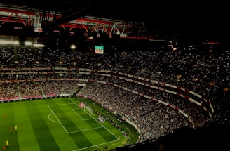 A large, illuminated stadium filled with a vast audience. The field is marked for soccer, with players actively participating in a match. The crowd is engaged, with numerous lights visible in the stands, possibly from phones. The stadium's roof structure is visible with red beams, and an electronic scoreboard shows a score of 0-0.