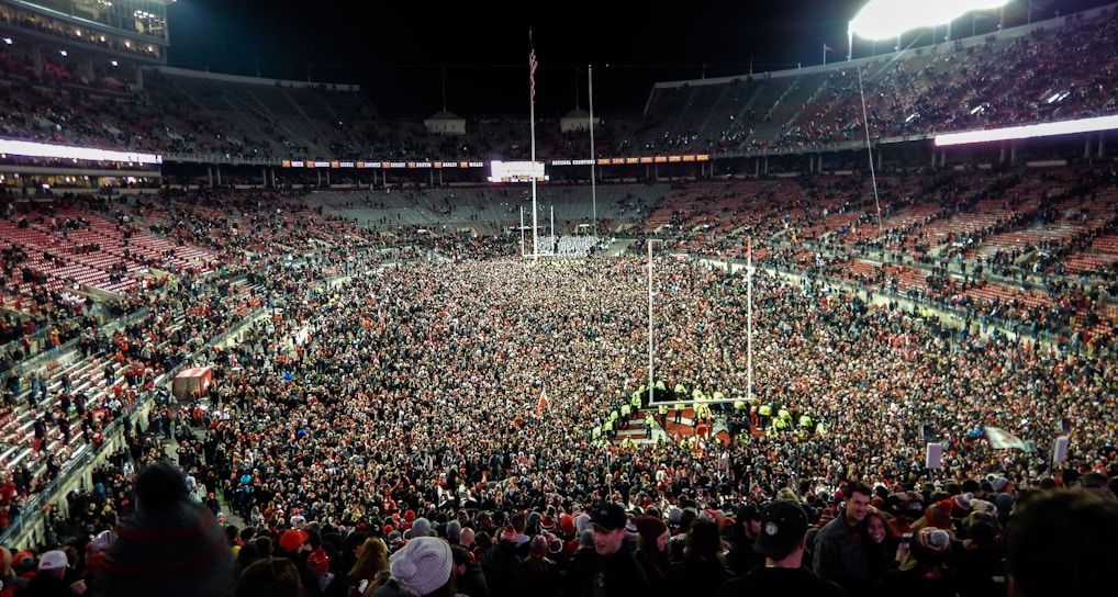 A massive crowd fills a sports stadium after an event, with people covering the field and the stands partially occupied. Bright stadium lights illuminate the scene, creating a vibrant atmosphere as individuals celebrate together.