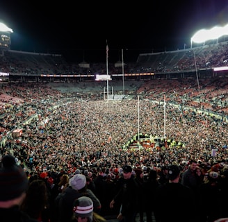 A massive crowd fills a sports stadium after an event, with people covering the field and the stands partially occupied. Bright stadium lights illuminate the scene, creating a vibrant atmosphere as individuals celebrate together.