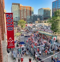 A vibrant outdoor scene featuring a large crowd of people gathered near a sports venue. A prominent red banner with '2018' and a sports logo hangs from a brick building. Nearby, there is a sign for a team store and souvenir shop. Surrounding the event are several modern high-rise buildings and a parking lot full of cars. Tall trees are visible in the background.