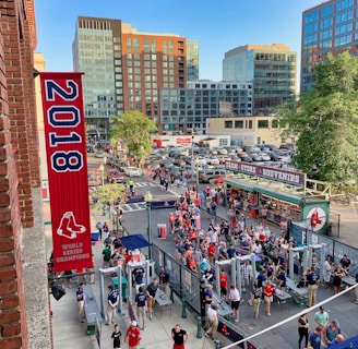 A vibrant outdoor scene featuring a large crowd of people gathered near a sports venue. A prominent red banner with '2018' and a sports logo hangs from a brick building. Nearby, there is a sign for a team store and souvenir shop. Surrounding the event are several modern high-rise buildings and a parking lot full of cars. Tall trees are visible in the background.