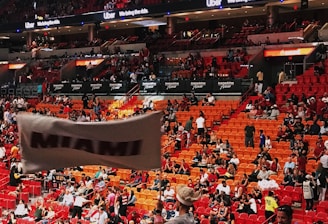 An indoor sports arena filled with a large crowd of spectators. People are sitting in tiered seating areas with red and orange tones dominating the seats. A person in the foreground is waving a large white flag with 'MIAMI' written on it. Numerous attendees wear basketball jerseys, and there is visible excitement and energy in the atmosphere. Advertisements for Uber are displayed prominently on digital boards near the ceiling.
