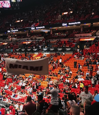 An indoor sports arena filled with a large crowd of spectators. People are sitting in tiered seating areas with red and orange tones dominating the seats. A person in the foreground is waving a large white flag with 'MIAMI' written on it. Numerous attendees wear basketball jerseys, and there is visible excitement and energy in the atmosphere. Advertisements for Uber are displayed prominently on digital boards near the ceiling.