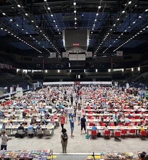 An indoor sports arena filled with rows of tables, each occupied by people engaged in competitive activities. The ceiling is high, with numerous lights illuminating the large space. The audience seating on either side is empty, and there are flags hanging from the upper level.