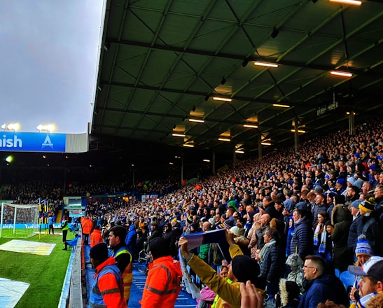 A crowded stadium filled with enthusiastic spectators. Many are standing and appear to be watching a sporting event with intensity. The stadium lighting is on, and the field is partially visible to the left. Some people are wearing team apparel, suggesting a sports team atmosphere. There are stewards wearing bright orange jackets along the edge of the field.
