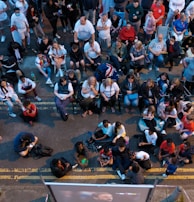 A large group of people gathered outdoors, watching a screen that is displaying a man. The crowd is diverse in age and appears interested and engaged, with many sitting closely together, some standing. Many are dressed casually, some in sports attire, and a few waving or holding flags. The event seems to have a communal, festive atmosphere.