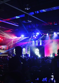 A dimly lit indoor arena filled with an audience. Bright stage lights in red and blue illuminate multiple large screens displaying graphics.