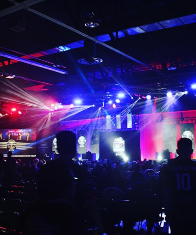 A dimly lit indoor arena filled with an audience. Bright stage lights in red and blue illuminate multiple large screens displaying graphics.