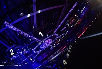 A large indoor arena with colorful and vibrant lighting, featuring a center stage surrounded by rigging and structures. Large numbered screens are visible, indicating marked sections from one to five. Various pieces of equipment and people are scattered around the venue.
