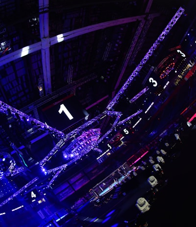 A large indoor arena with colorful and vibrant lighting, featuring a center stage surrounded by rigging and structures. Large numbered screens are visible, indicating marked sections from one to five. Various pieces of equipment and people are scattered around the venue.