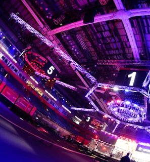An indoor arena with vibrant purple and blue lighting, featuring large numbered screens suspended from the ceiling. The venue has a high ceiling with intricate metal truss structures and hanging lights, creating a futuristic and energetic atmosphere.
