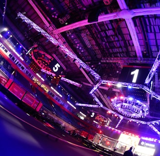 An indoor arena with vibrant purple and blue lighting, featuring large numbered screens suspended from the ceiling. The venue has a high ceiling with intricate metal truss structures and hanging lights, creating a futuristic and energetic atmosphere.