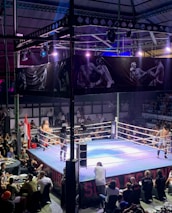 A crowded indoor arena with a boxing ring at the center, surrounded by an audience seated at tables. The scene is illuminated by overhead lights, casting a vibrant glow over the ring where two fighters are preparing for a match. Spectators are engaged, capturing the event with cameras and enjoying the lively atmosphere.