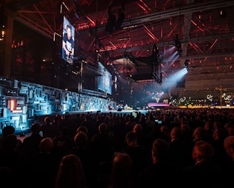A large indoor event with a stage featuring bright screens and spotlights, attended by a substantial crowd. The venue has a high ceiling with industrial beams and red lighting accents. The atmosphere resembles a concert or a conference with a focus on visual effects.