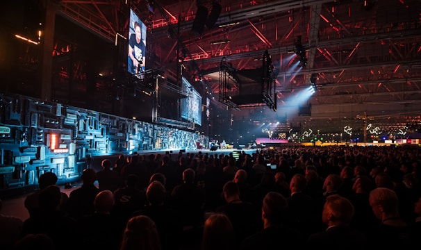 A large indoor event with a stage featuring bright screens and spotlights, attended by a substantial crowd. The venue has a high ceiling with industrial beams and red lighting accents. The atmosphere resembles a concert or a conference with a focus on visual effects.